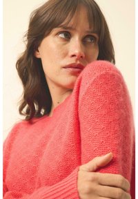 Heller korallfarbener Strickpullover mit gerippten Bündchen und einer weichen, strukturierten Oberfläche. Das Garn wirkt flauschig und verleiht einen gemütlichen Look. Keine sichtbaren Muster.