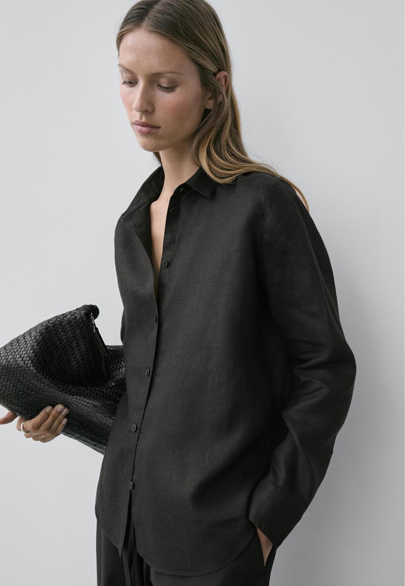 Vrouw met lang lichtbruin haar draagt een zwart blouse met knopen, houdt een grote zwarte geweven clutch vast en kijkt omlaag tegen een grijze achtergrond.