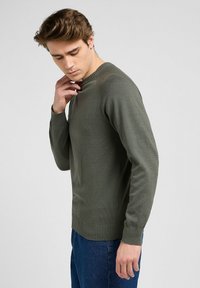 Suéter de punto verde oliva con cuello redondo, puños ribeteados y dobladillo. Presenta una textura suave y una silueta ajustada. Detalle sutil de la marca en la manga.