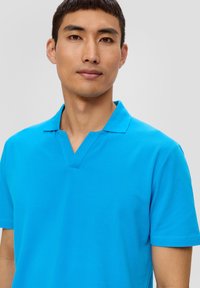 Helles blaues Polohemd mit V-Ausschnitt, kurzen Ärmeln und strukturierter Stoffqualität. Minimale Nähte und klare Linien sind durchgängig vorhanden.