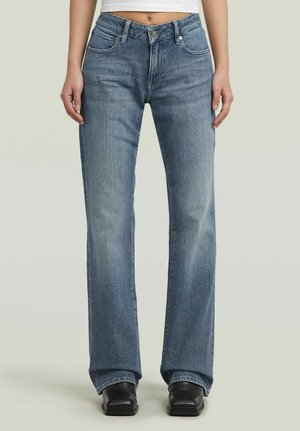 Vrouw draagt high-waisted, rechte blauwe spijkerbroek met steekzakken aan de voorkant en zwarte schoenen met vierkante neus tegen een effen achtergrond.