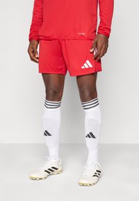 Κόκκινη αθλητική φανέλα και σορτς με το λογότυπο της Adidas, λευκές κάλτσες με μαύρες ρίγες και λευκά ποδοσφαιρικά παπούτσια με μαύρες λεπτομέρειες.