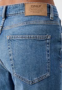Primer plano de una persona que lleva unos vaqueros de mezclilla azul, mostrando el bolsillo trasero y la etiqueta de marca de cuero encima de la cinturilla.