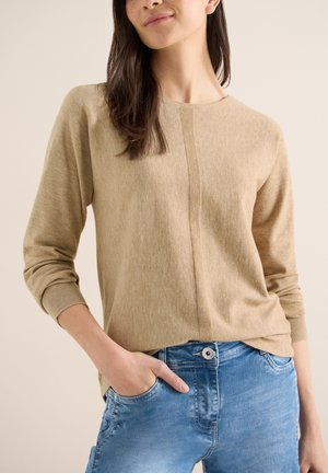 Femme portant un pull beige à manches longues et un jean bleu, une main dans la poche, debout devant un fond clair uni.