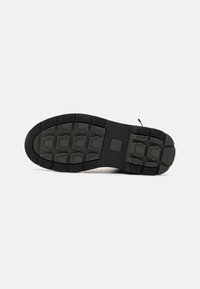 Semelle de chaussure noire avec des motifs de bande de roulement rectangulaires et hexagonaux et un petit logo carré au centre, sur un fond clair uni.