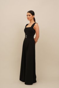 Melna kombinezons ar kvadrātveida dekoltē, platām lencēm, jostu viduklī un platām biksēm ar krokām, kas izgatavots no gludas, teksturētas auduma.