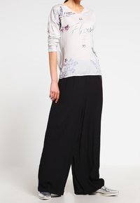 Camicia a maniche lunghe di colore grigio chiaro con design floreale e testo, abbinata a pantaloni neri larghi. Il modello indossa scarpe argentate.