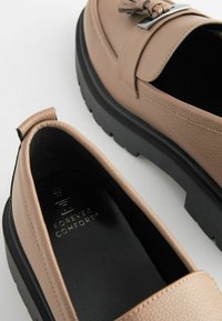 Mocasines de cuero beige con un acabado texturizado y una suela gruesa negra. Acento de hardware plateado en la parte superior. Logo de la marca en el interior.