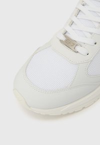 Fehér sportcipő texturált hálós felsőrésszel, sima szintetikus díszítésekkel, lapos fűzőkkel és ezüst színű logóval a nyelvén.