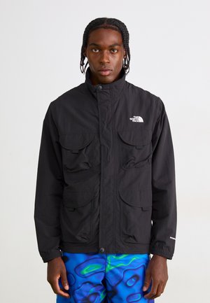 Young man with braided hair wearing a black The North Face jacket and blue patterned pants, standing against a plain white background.