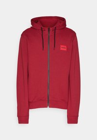 Červená zipová mikina vyrobená z měkké tkaniny, s kapucí, černými šňůrkami a malou obdélníkovou nášivkou s logem na levé stran�ě hrudi.