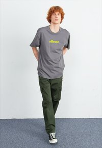 Grijze t-shirt met groene "ellesse" logo, gecombineerd met olijfgroene cargo broek en zwarte hoge sneakers. Model staat met de handen achter de rug.