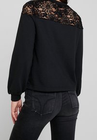 Sudadera negra con superposición de encaje floral en los hombros, con mangas largas abullonadas y puños de canalé, combinada con jeans negros.