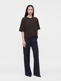 Femme aux longs cheveux lisses portant un t-shirt noir oversize, un pantalon large bleu marine et des chaussures noires pointues, debout devant un fond uni.