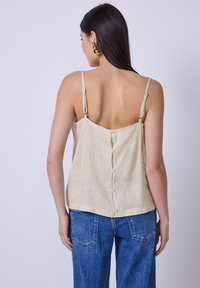 Camisola di lino beige con spalline sottili e regolabili, caratterizzata da una chiusura a bottone sul retro, abbinata a jeans in denim blu a vita alta.