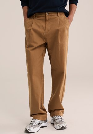 Mann trägt lockere braune Hose und weiße Laufschuhe, steht mit Händen in den Taschen vor einfachem Hintergrund.