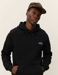 Młody mężczyzna w czarnej bluzie z kapturem z małym białym logo i brązowej czapce w deseń, stojący na tle jasnego, jednolitego tła.