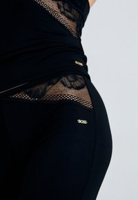 Mustan urheiluvaatteen yläosa, jossa on pitsi- ja verkkokudon yksityiskohtia sekä kultaisia logoesittelyjä. Istuva muotoilu, jossa on teksturoitu kangas, korostaen vartalon muotoja.