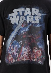 Černé bavlněné tričko s barevnou grafikou "Star Wars", zobrazující ikonické postavy a prvky na pozadí hvězdné oblohy.