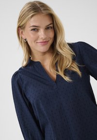 Blusa blu navy con scollo a V, caratterizzata da sottili pois testurizzati e maniche lunghe. Tessuto morbido con dettagli arricciati sulle spalle e una vestibilità comoda.