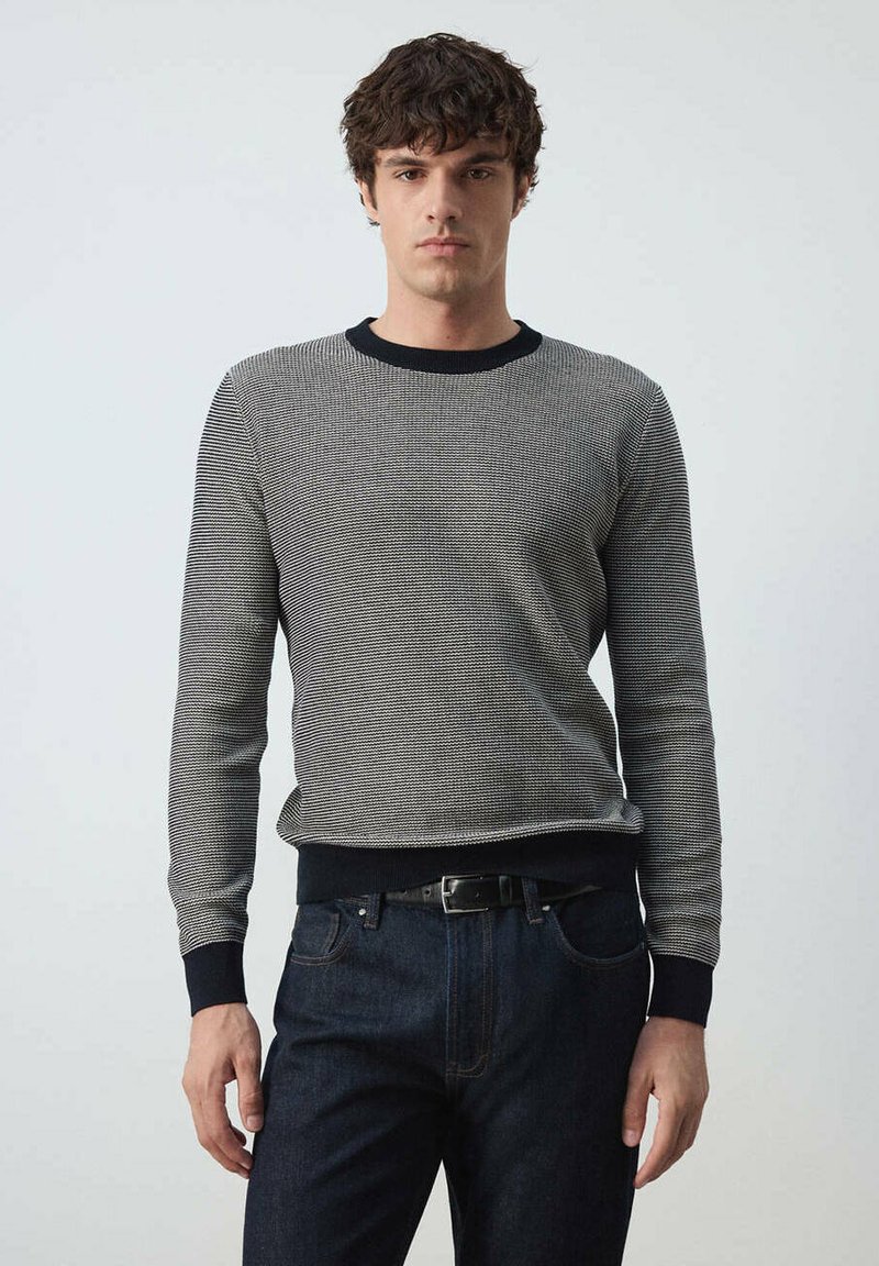 Jeune homme portant un pull à manches longues à rayures noires et blanches avec des poignets noirs et un jean bleu foncé, debout devant un fond uni.