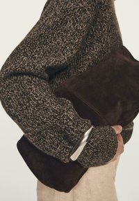 Osoba w grubej brązowej swetrze w wzory, trzymająca dużą ciemnoczekoladową zamszową kopertówkę na jasnym tle.