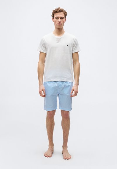 Νέος άντρας όρθιος ξυπόλητος, φορώντας λευκό κοντομάνικο πουκάμισο και μπλε-λευκές ριγέ κοντές βερμούδες, μπροστά σε απλό λευκό φόντο.