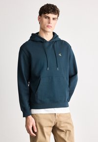 Felpa hoodie color verde scuro con tasca frontale, coulisse e un piccolo logo giallo sul petto. Realizzata in tessuto morbido con polsini a costine.