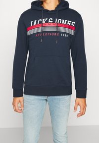 Sudadera azul marino con bolsillo canguro, capucha con cordón y franjas en rojo y blanco que presentan el texto "JACK & JONES".