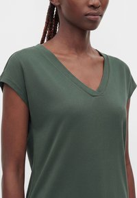 Femme portant un haut vert foncé à col en V à manches courtes, montrant les épaules et le haut de la poitrine sur un fond uni.