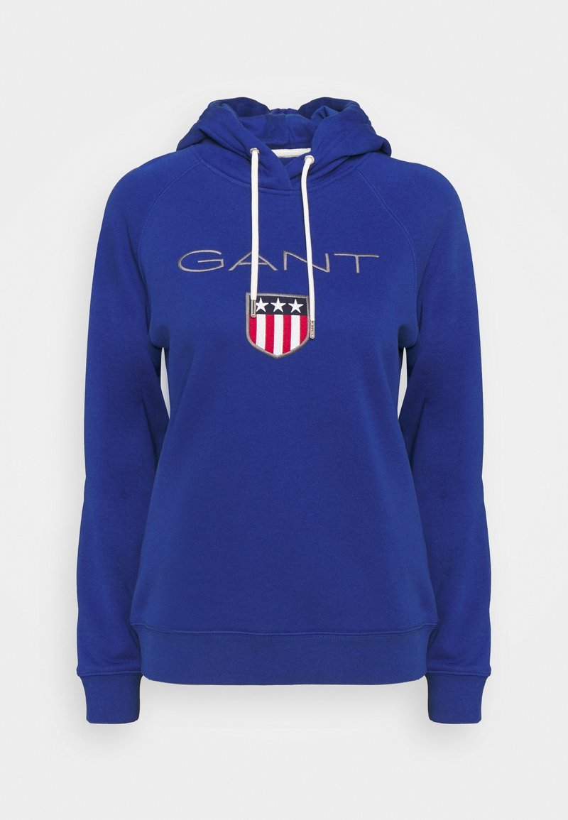 Sweat à capuche bleu avec cordons blancs, texte du logo GANT et un emblème en forme de bouclier avec des étoiles et des rayures sur la poitrine.
