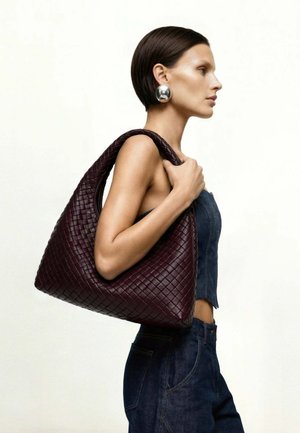 Femme en tenue en jean portant un grand sac à main en cuir tressé marron foncé sur l'épaule, portant de grandes boucles d'oreilles rondes argentées, tournée vers la droite.