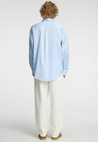 Jasnoniebieska koszula na guziki z pionowymi paskami, długimi rękawami, zaokrąglonym dołem i luźnym krojem, połączona z luźnymi białymi spodniami.