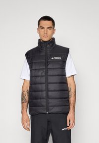 Black padded vest with high neck, front zipper, and logo detail. Worn over a white shirt, paired with dark pants. Features textured fabric.