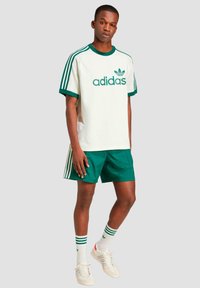 Žalias ir baltas Adidas sportinis komplektas, kuriame yra trumpomis rankovėmis marškiniai su tinklinėmis rankovėmis, derančios šortai ir dryžuotos socės.
