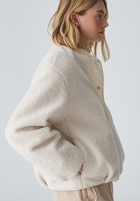 Veste en polaire crème avec une coupe décontractée, des poignets côtelés, une fermeture à boutons-pression et des poches latérales. Texture douce et design minimal.
