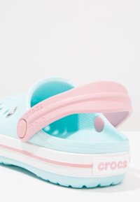 Aqua blue Crocs with a pink strap, featuring a perforated design, white midsole, and raised logo on the heel. Lightweight, slip-on style.