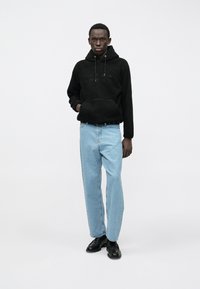 Νεαρός άνδρας με μαύρη φλις φούτερ, ανοιχτό μπλε τζιν και μαύρα παπούτσια που στέκεται μπροστά σε λευκό φόντο.