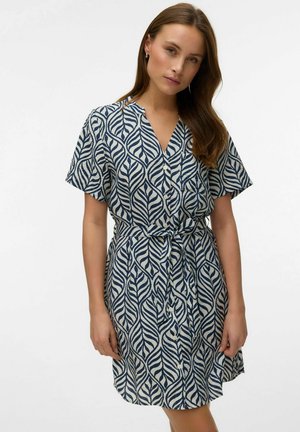 Kvinde iført en knap-op kjole med korte ærmer, navy og hvidt bladmønster, bundet ved taljen, stående mod en ensfarvet baggrund.