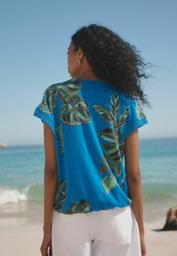 Haut bleu à manches courtes avec un motif de feuilles tropicales. Tissu léger avec une bordure en dentelle au niveau des manches. Porté avec un pantalon blanc devant un fond de plage.