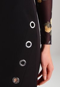 Robe noire avec des œillets en métal argenté sur le côté, accompagnée d'une douce manche longue en tissu floral transparent. La texture semble lisse et ajustée.