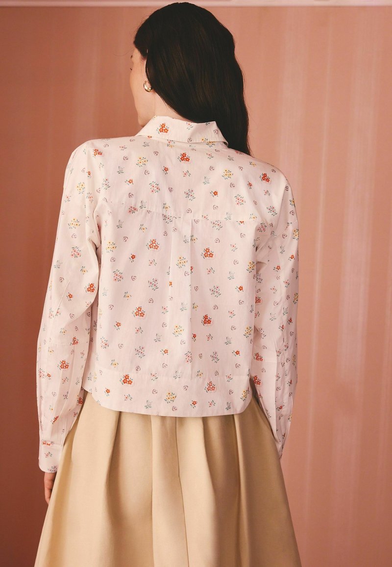 Žena s dlhými tmavými vlasmi má na sebe bielu kvetovanú blúzku s dlhými rukávmi a béžovú plisovanú sukňu, stojaca pred broskyňovým pozadím.