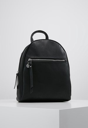 Sac à dos en cuir noir avec poche zippée à l'avant et poignée supérieure, posé sur une surface blanche devant un fond gris uni.