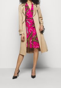 Dlouhý béžový trenčkot přes růžové šaty s vzorem řetězu. Černé vysoké podpatky. Trenčkot má pásek a volný střih.