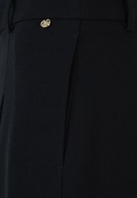 Pantaloni neri su misura realizzati in un tessuto liscio e testurizzato, con un unico bottone dorato visibile e dettagli con pieghe frontali.