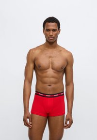 Κόκκινα ανδρικά εσώρουχα τύπου μποξέρ με μαύρη μέση που φέρει το λογότυπο "HUGO", παρουσιάζοντας ένα λείο μείγμα βαμβακιού και εφαρμοστή γραμμή.