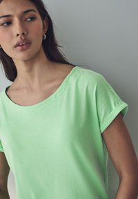 T-shirt verde chiaro a maniche corte con scollatura ampia, realizzato in tessuto morbido; presenta maniche arrotolate e una vestibilità rilassata.