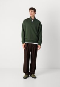 Felpa verde con zip e colletto alto, abbinata a pantaloni larghi marroni e calzature verdi. Tessuto morbido con una vestibilità rilassata.