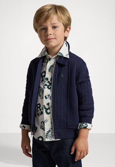 Casaco azul-marinho acolchoado com fecho éclair sobre uma camisa branca com padrões verdes, combinado com jeans escuros. Mangas curtas, punhos canelados.
