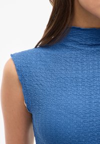 Blått texturerat plagg med hög hals, korta ärmar och ojämna kanter. Tyget har ett subtilt, vävt mönster.